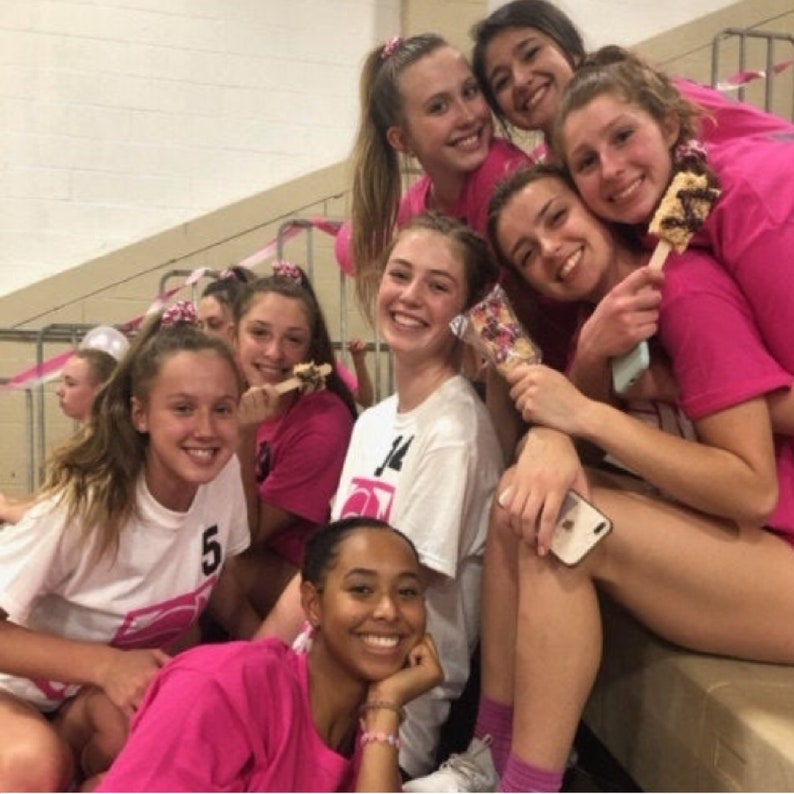 May include: A group of young women wearing pink shirts and holding popsicles. Some of the women are wearing hair bows. The women are smiling and looking at the camera.