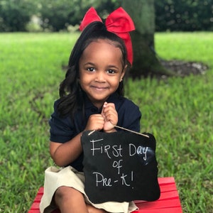 May include: A young child with dark hair and a large red bow smiles, holding a chalkboard sign that reads "First Day of Pre-K!". She wears a navy blue polo shirt and khaki skirt, sitting on a red bench against a green grassy background.