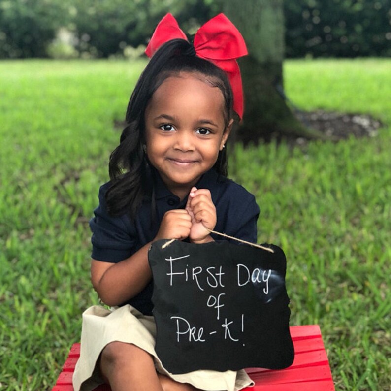 May include: A young child with dark hair and a large red bow smiles, holding a chalkboard sign that reads "First Day of Pre-K!". She wears a navy blue polo shirt and khaki skirt, sitting on a red bench against a green grassy background.