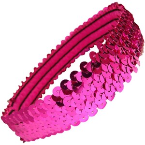 May include: A bright pink sequined headband. The headband features a row of shiny, round sequins attached to a pink elastic band. The elastic band has black stripes. The headband is designed to be worn around the head.