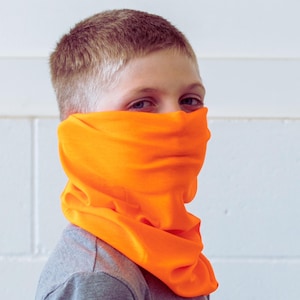 May include: Bright orange neck gaiter worn around the neck and covering the lower face. The gaiter is made of a soft, flexible material. The person is wearing a gray shirt. The background is a white wall.