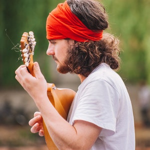 May include: A person with a red headband and white t-shirt playing a wooden acoustic guitar. The guitar has silver tuning pegs. The background is blurred green foliage.
