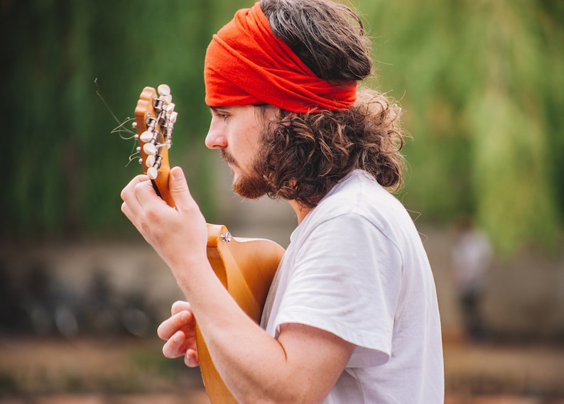 May include: A person with a red headband and white t-shirt playing a wooden acoustic guitar. The guitar has silver tuning pegs. The background is blurred green foliage.
