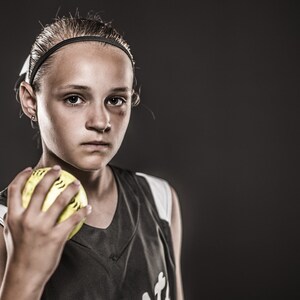 May include: A young person wearing a black and white jersey holds a yellow softball in their hand. They are looking directly at the camera with a serious expression.