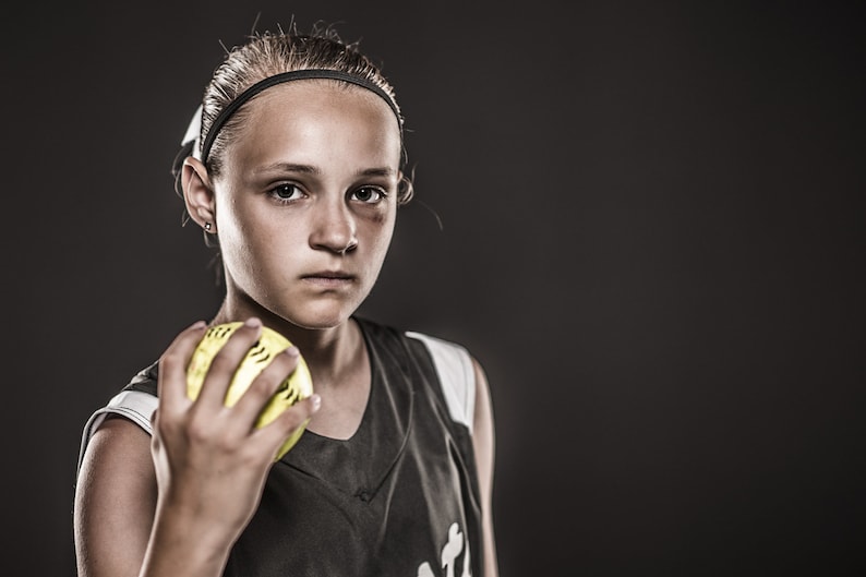 May include: A young person wearing a black and white jersey holds a yellow softball in their hand. They are looking directly at the camera with a serious expression.