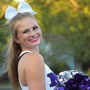 May include: A smiling cheerleader wearing a white top with a purple "M" logo and a white bow in her hair. She holds purple and silver pom-poms, with a blurred green background.