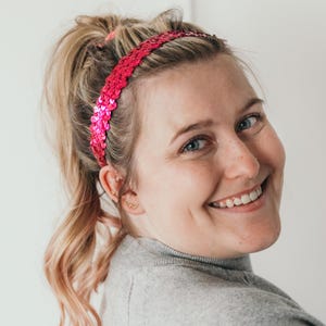 May include: A bright pink sequin headband. The headband is made of small, shiny sequins and is worn across the forehead. The hair is blonde and pulled back in a ponytail. The person is wearing a gray turtleneck.