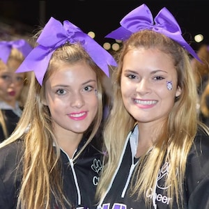 May include: Two young women wearing black cheerleading uniforms with purple bows in their hair. The uniforms have the word "Cheer" printed on them.