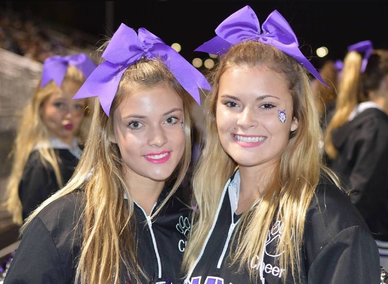 May include: Two young women wearing black cheerleading uniforms with purple bows in their hair. The uniforms have the word "Cheer" printed on them.
