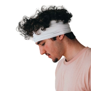May include: A person wearing a white headband and a light pink t-shirt. The person has dark curly hair and a short beard. The background is plain white.