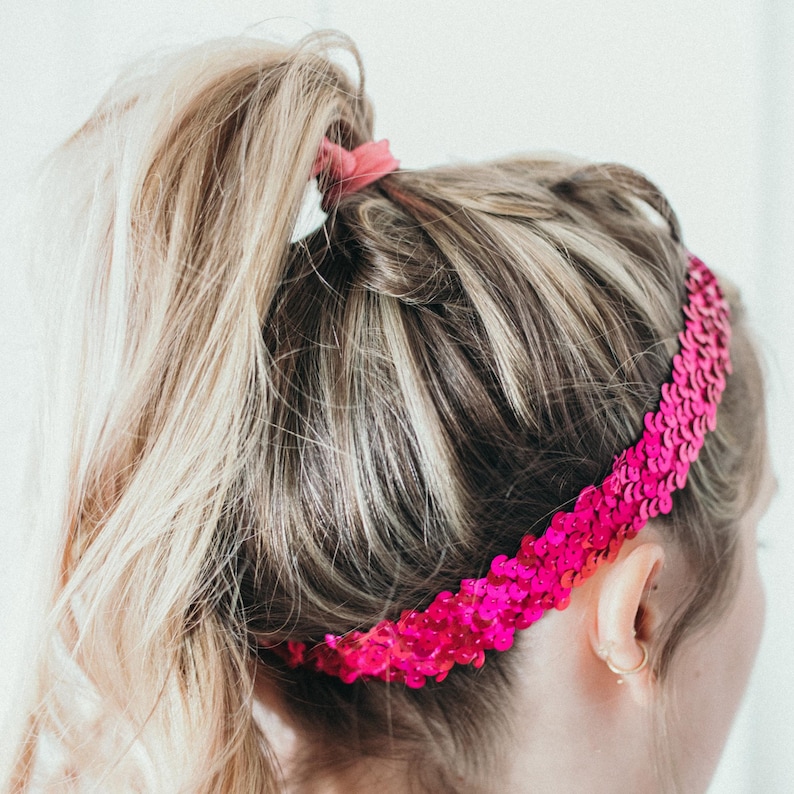 May include: A bright pink sequin headband worn on the head. The headband is made of small, shiny sequins and is wrapped around the head. The hair is blonde and pulled back in a ponytail.