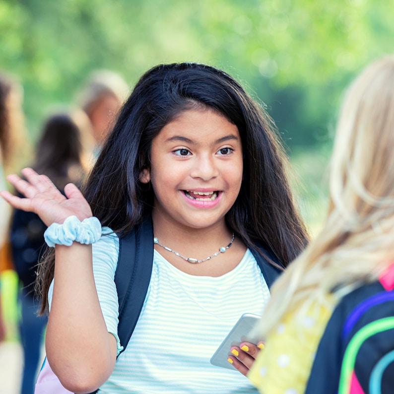 Puede incluir: Una joven con el pelo largo y oscuro sonr&iacute;e y saluda mientras sostiene un tel&eacute;fono m&oacute;vil. Lleva una camisa a rayas azul y blanca, una mochila negra y una goma de pelo azul en la mu&ntilde;eca.