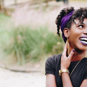 May include: A person wearing a black top and a purple head wrap, with gold earrings and a gold watch. The person is smiling and looking upwards. The background is blurred with green and tan tones.