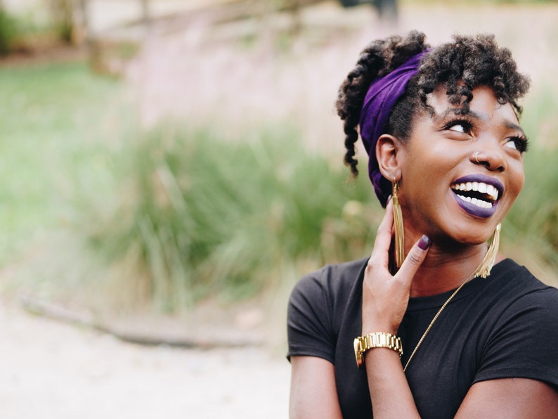 May include: A person wearing a black top and a purple head wrap, with gold earrings and a gold watch. The person is smiling and looking upwards. The background is blurred with green and tan tones.