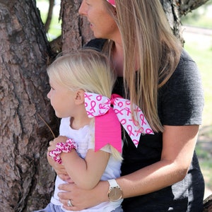 May include: A woman and a young girl are wearing pink ribbon headbands and bows. The woman is wearing a gray shirt and the girl is wearing a white shirt and a gray skirt. They are standing in front of a tree.