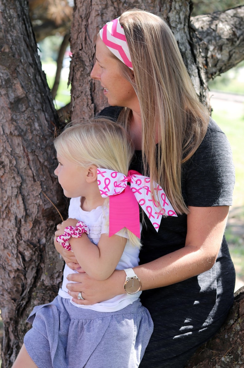 May include: A woman and a young girl are wearing pink ribbon headbands and bows. The woman is wearing a gray shirt and the girl is wearing a white shirt and a gray skirt. They are standing in front of a tree.