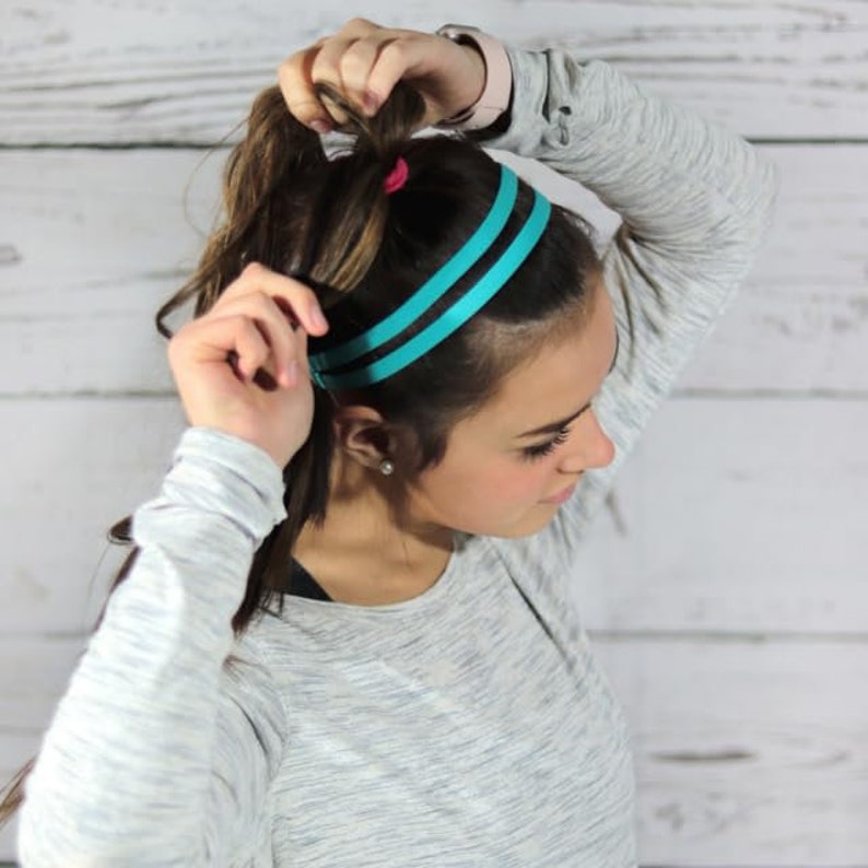 May include: A woman with brown hair is wearing a teal headband with two stripes. She is wearing a gray long-sleeved shirt.