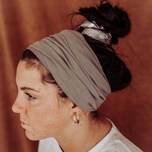 May include: A gray fabric headband is wrapped around the head, with a silver hair accessory at the back. The person is wearing gold hoop earrings and a white shirt. The background is a solid brown color.
