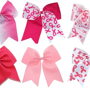 May include: Six pink and white cheer bows with different designs. The bows feature pink ribbons with white text and pink ribbons with white and pink ribbon designs. The bows are tied with a white ribbon.