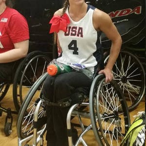 May include: A woman wearing a white and blue basketball jersey with the word "USA" and the number "4" on the front, sits in a wheelchair. She is smiling and has a red bow in her hair. The wheelchair has two large wheels and two smaller wheels.