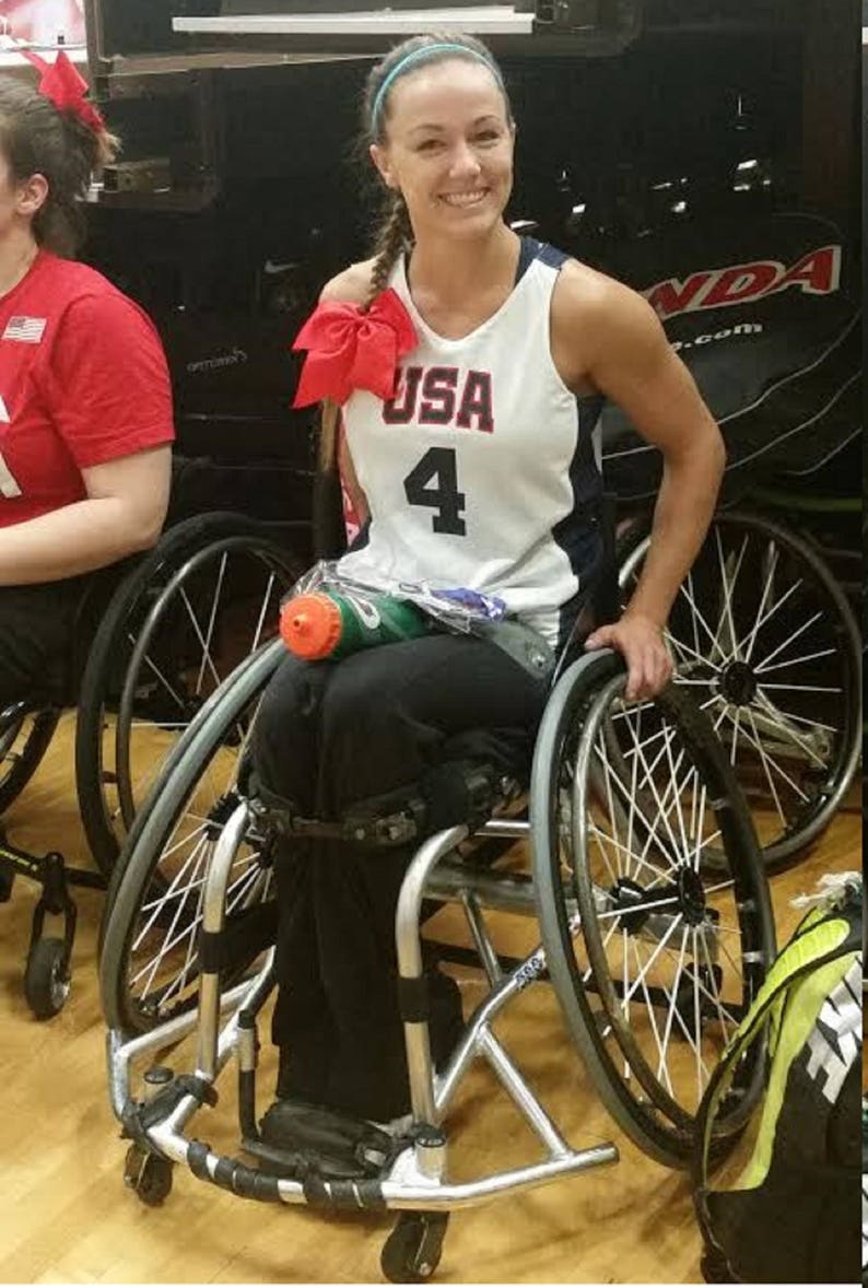 May include: A woman wearing a white and blue basketball jersey with the word "USA" and the number "4" on the front, sits in a wheelchair. She is smiling and has a red bow in her hair. The wheelchair has two large wheels and two smaller wheels.