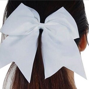 May include: A large, white cheerleading bow. The bow is made of a smooth, satin-like material and is tied in a classic bow shape. The bow is positioned on the back of a person's head, with their dark hair visible.