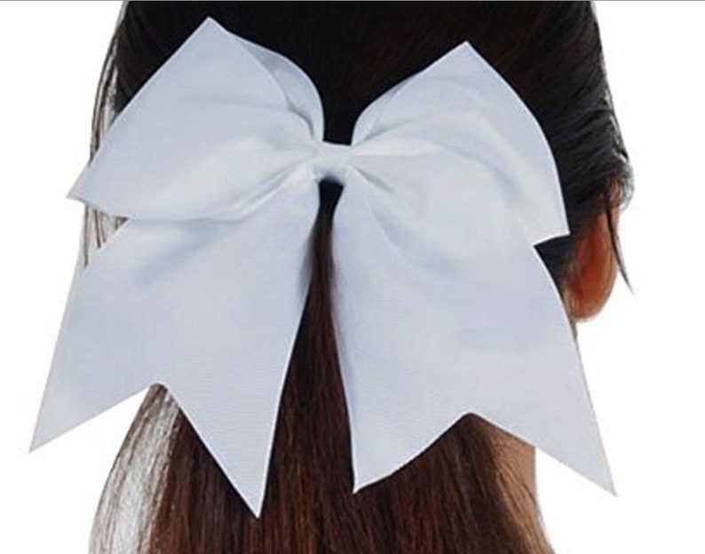 May include: A large, white cheerleading bow. The bow is made of a smooth, satin-like material and is tied in a classic bow shape. The bow is positioned on the back of a person's head, with their dark hair visible.