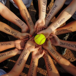 May include: A group of people with their arms linked together, each arm has a motivational word written on it. The words are: Get Dirty, Passion, Believe, Enthusiasm, Attitude, Teamwork, Confidence, Play Hard, Fight, Determination, Relax, Stay Positive, Go Hard, Integrity. A yellow softball is in the center of the circle.