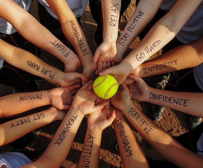 May include: A group of people with their arms linked together, each arm has a motivational word written on it. The words are: Get Dirty, Passion, Believe, Enthusiasm, Attitude, Teamwork, Confidence, Play Hard, Fight, Determination, Relax, Stay Positive, Go Hard, Integrity. A yellow softball is in the center of the circle.