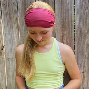 K&ouml;nnte beinhalten: Eine junge Person mit langen blonden Haaren, die ein dunkelrotes Stirnband und ein hellgelbes geripptes Tanktop tr&auml;gt.