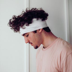 May include: A person wearing a light pink t-shirt and a white headband. The person has dark curly hair and a short beard. The background is a plain white wall.