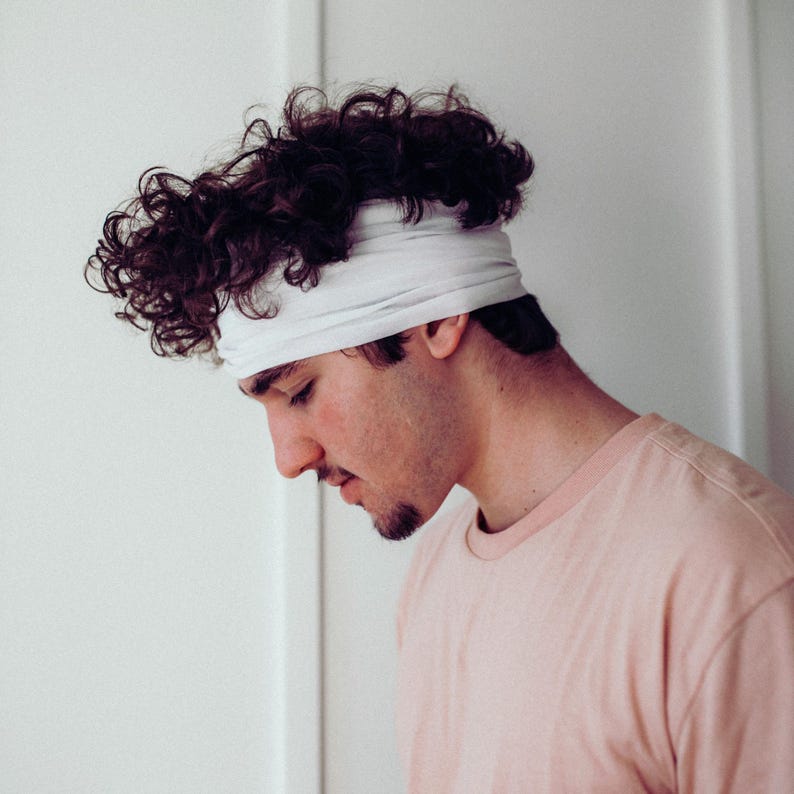 May include: A person wearing a light pink t-shirt and a white headband. The person has dark curly hair and a short beard. The background is a plain white wall.