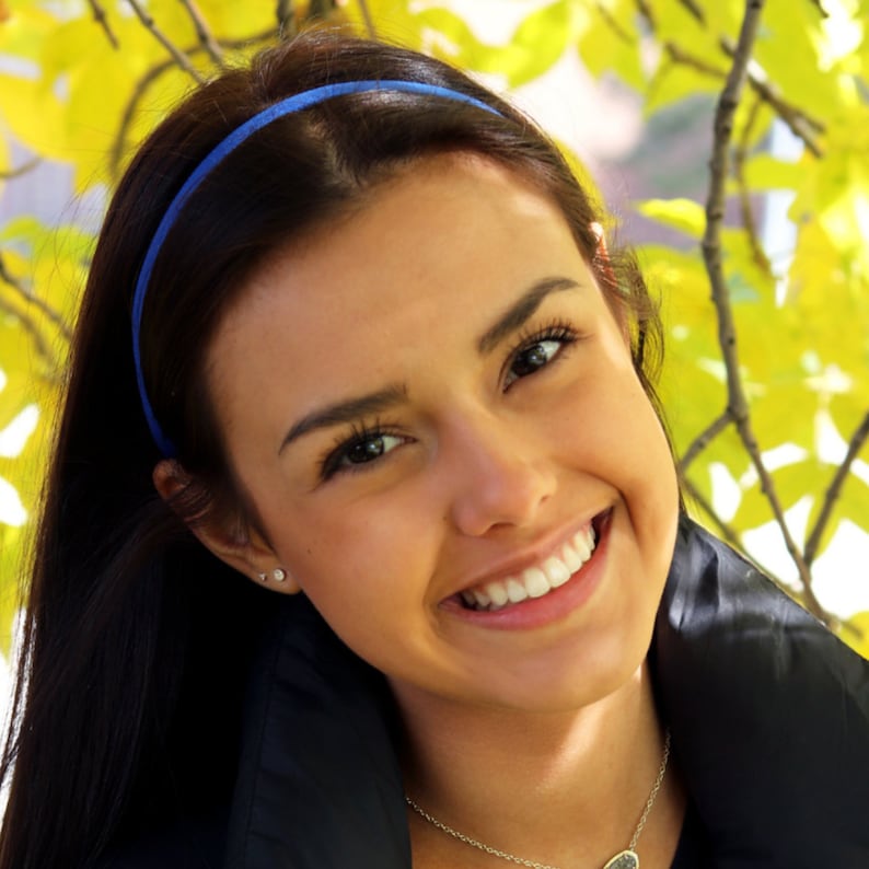 May include: A young woman with long dark hair is smiling. She is wearing a blue headband and a black jacket. The background is a blurry image of yellow leaves.