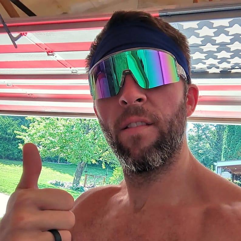 K&ouml;nnte beinhalten: Ein Mann mit einem blauen Stirnband und regenbogenfarbenen gr&uuml;nen Sonnenbrillen mit wei&szlig;em Rahmen. Er zeigt den Daumen nach oben und hat einen Bart.