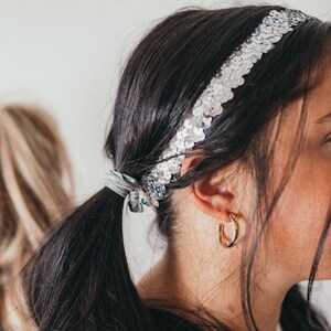 May include: A woman with dark hair wearing a silver sequin headband and gold hoop earrings. Her hair is pulled back in a ponytail, secured with a silver scrunchie. The headband is adorned with small, reflective sequins.