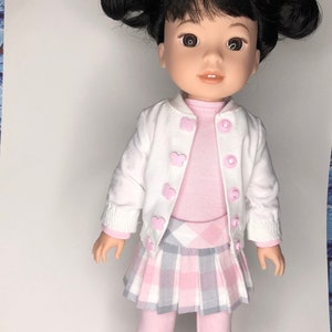 May include: A doll with black hair in pigtails wearing a white cardigan with pink butterfly buttons, a pink long-sleeved shirt, a plaid skirt with pink and gray squares, pink tights, and black shoes.