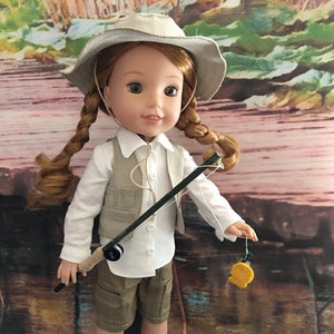 Può includere: Una bambola con un cappello beige, una camicia bianca e pantaloncini color kaki tiene una canna da pesca con un'esca. La bambola ha i capelli rossi intrecciati e gli occhi marroni. Lo sfondo è un dipinto ad acquerello di un ruscello.