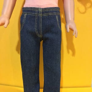 May include: A pair of blue denim jeans with yellow stitching. The jeans are made for a doll.