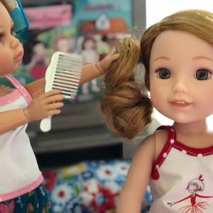 May include: Two dolls, one with pink and white polka dot tank top and blue and green patterned pants, is combing the hair of the other doll who is wearing a white tank top with red trim and white polka dots, and a red and white ballerina graphic.