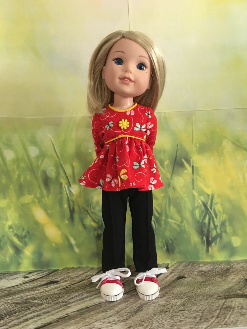 Op de afbeelding: Een pop met blond haar die een rood en geel bloempatroon top, een zwarte broek en rode sneakers draagt.