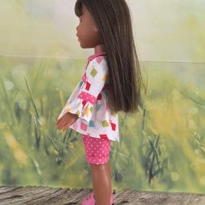 Op de afbeelding: Een pop met lang bruin haar die een wit en roze jurkje draagt met een geometrisch patroon en roze stippen shorts. De pop draagt roze schoenen.