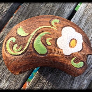 Puede incluir: Un plato de cerámica marrón con un diseño floral verde y blanco. El plato tiene forma de media luna y tiene un borde elevado.