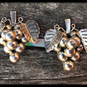 Op de afbeelding: Een paar zilveren druiventros hangers met gouden bladeren accenten. De druiven zijn rond en hebben een glanzende afwerking. De bladeren zijn getextureerd en hebben een licht geoxideerde uitstraling.