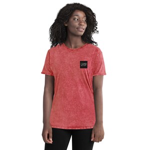 Op de afbeelding: Een rood, zuurgewassen T-shirt met een zwarte vierkante patch op de linkerborst. De patch heeft witte tekst die "CREATIVE" en "The Hustle is Real" luidt.
