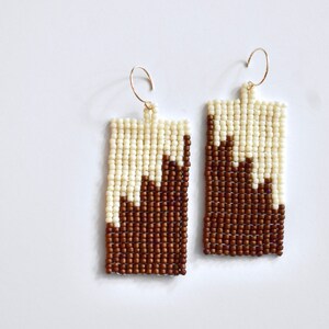 May include: A pair of brown and white beaded earrings with a geometric design. The earrings are rectangular in shape and have a gold hook.