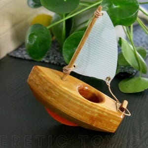 Könnte beinhalten: Ein Holzspielzeug-Segelboot mit hellem Segel und braunem Holzrumpf. Das Boot hat ein kleines Loch in der Mitte und einen orangefarbenen Sockel. Der Text "HERETIC TOYS CO" ist unten sichtbar.