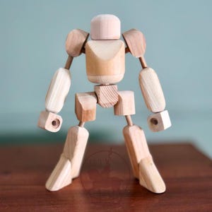 May include: A wooden artist's mannequin with articulated joints. The figure is made of light-colored wood and has a rounded head, torso, arms, and legs. The joints allow for various poses, making it suitable for drawing or display.