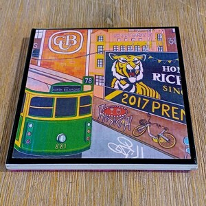 May include: A square coaster featuring a colorful illustration of a green North Richmond tram numbered 881, a tiger graphic, a bicycle, and a cafe table. The artwork includes text such as GB, Bryant & May, and 2017 PREM, set against a city street scene.