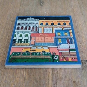 May include: A square ceramic tile featuring a colorful illustration of a city street scene. The image depicts buildings including Piedmonte's Supermarket and a movie theater, with a yellow car parked in front and stylized green foliage.