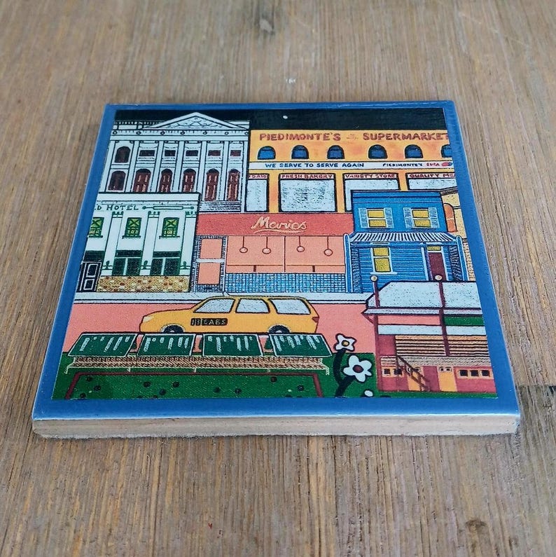 May include: A square ceramic tile featuring a colorful illustration of a city street scene. The image depicts buildings including Piedmonte's Supermarket and a movie theater, with a yellow car parked in front and stylized green foliage.